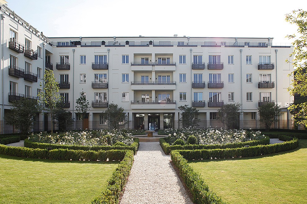 Gerader Kiesweg zwischen Rasen und Buchsbaumhecken im Innenhof, am Ende die helle Fassade eines Wohnhauses