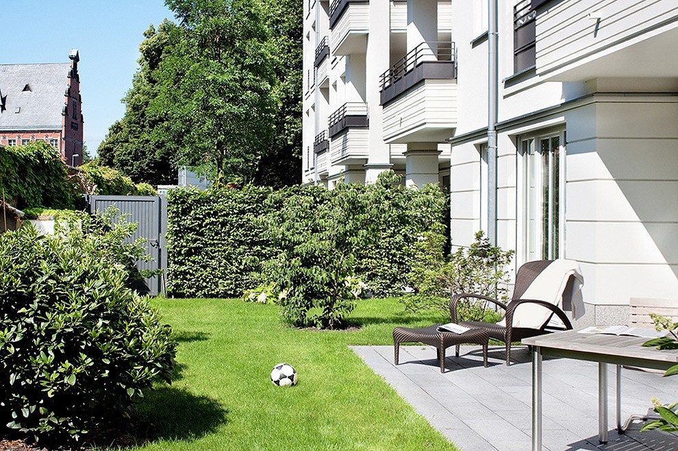 Privatgarten mit Rasen, Hecken, Liegestuhl und Terrasse vor einem weißen Wohnhaus, links ein Backsteinturm