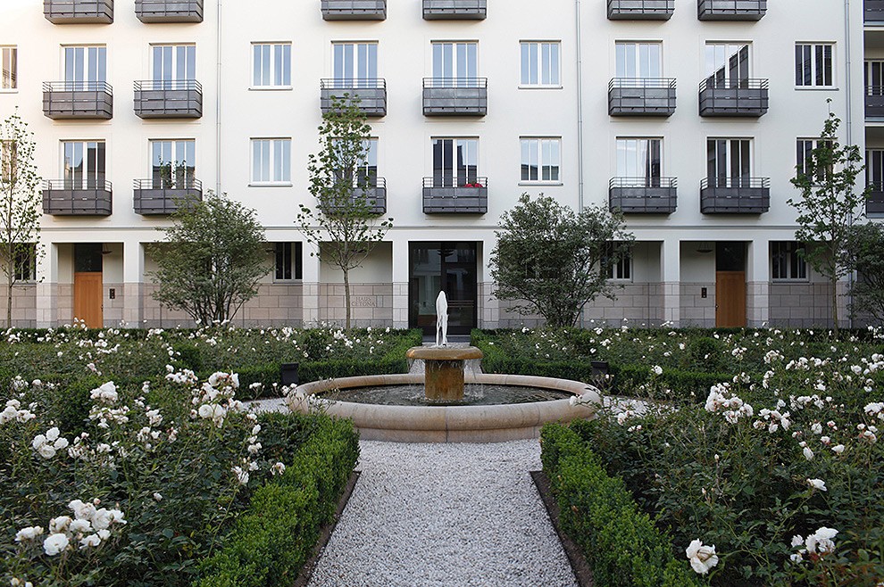 Runder Brunnen inmitten weißer Rosenbeete und Buchsbaumhecken vor einer hellen Wohnhausfassade
