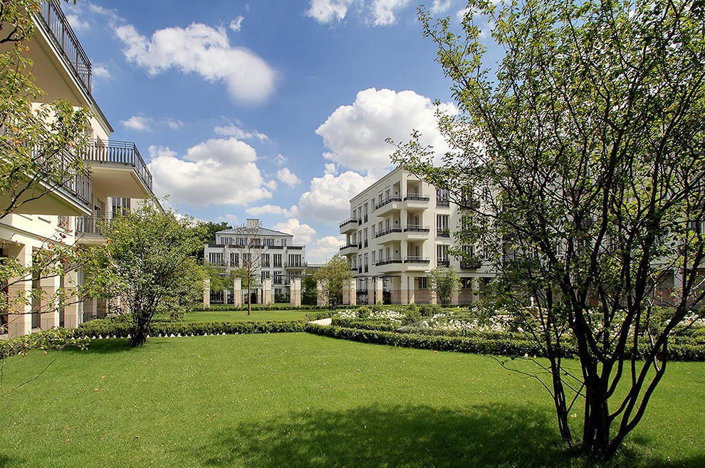 Große Rasenfläche im Innenhof mit Baum im Vordergrund, dahinter helle Wohnhäuser unter blauem Himmel