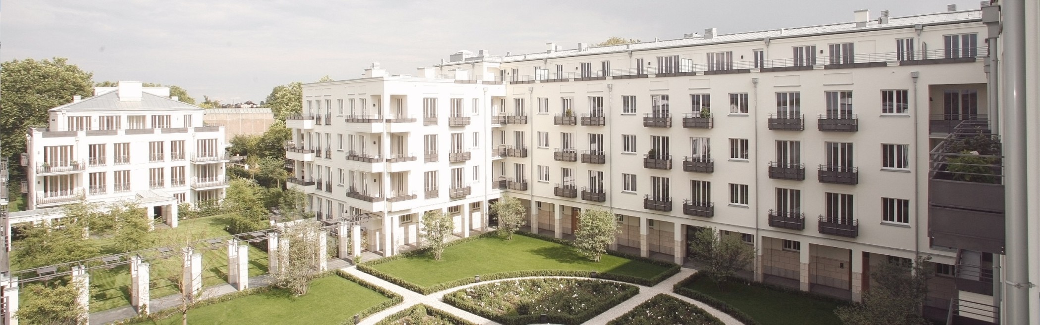 Erhöhter Blick auf den begrünten Innenhof mit Wegen und Pergola vor langen hellen Wohnhausfassaden