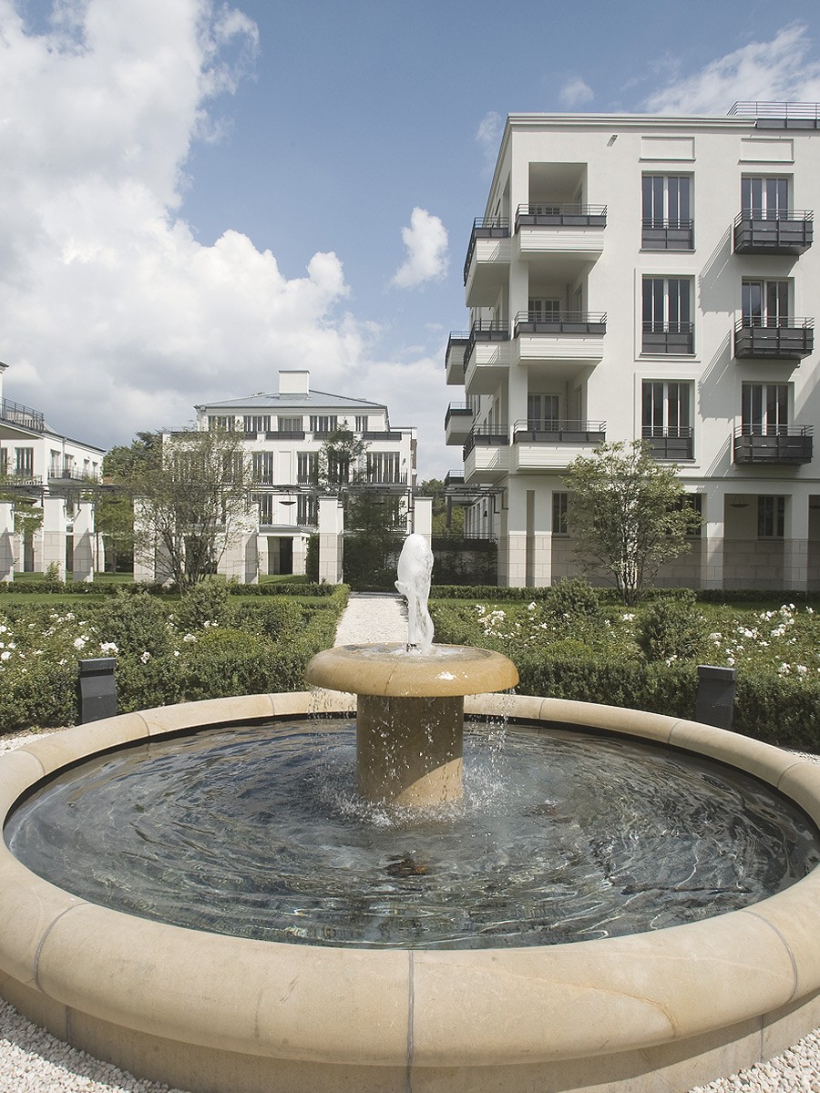 Runder Brunnen aus hellem Stein mit Fontäne im Innenhof, dahinter Gartenanlage und weiße Wohnhäuser