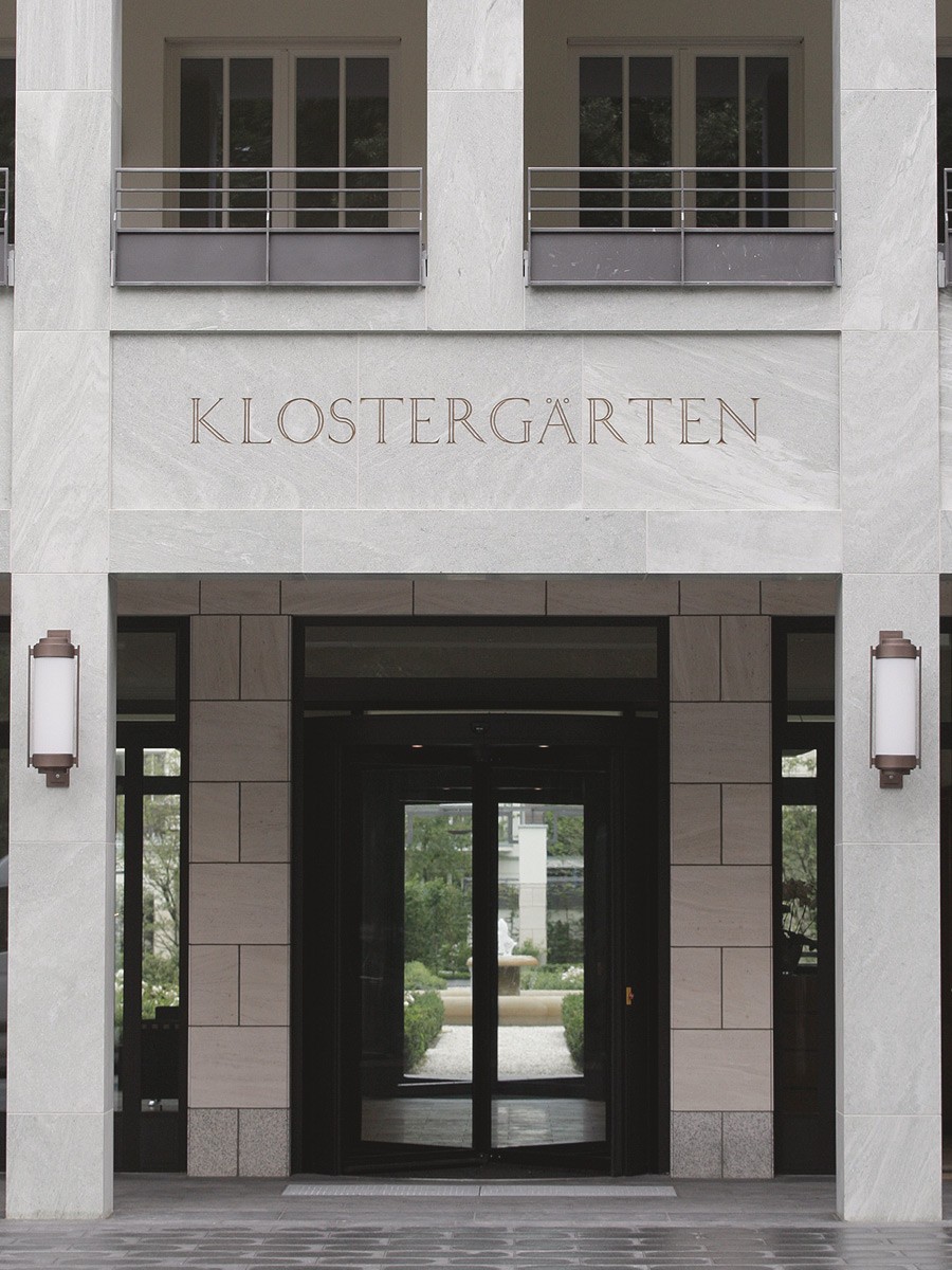 Eingangstür aus dunklem Glas unter der in Stein gemeißelten Inschrift Klostergärten, flankiert von Wandleuchten